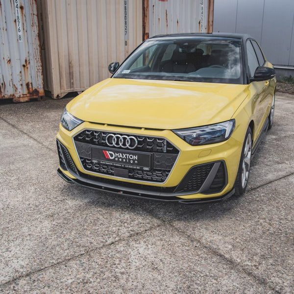 A yellow car is parked on a concrete surface near rusted shipping containers. The front license plate reads "MAXTON design."