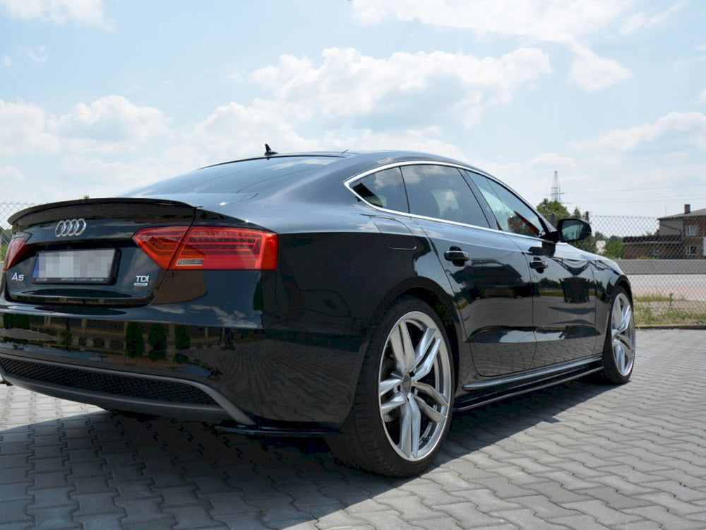 A black Audi A5 parked on a paved area under a partly cloudy sky. The car's sleek design and silver alloy wheels are prominent. Text includes "A5" and "TDI."