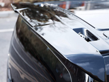 A sleek, glossy car spoiler is installed on the rear of a black vehicle, reflecting trees and sky, situated in an outdoor parking area.
