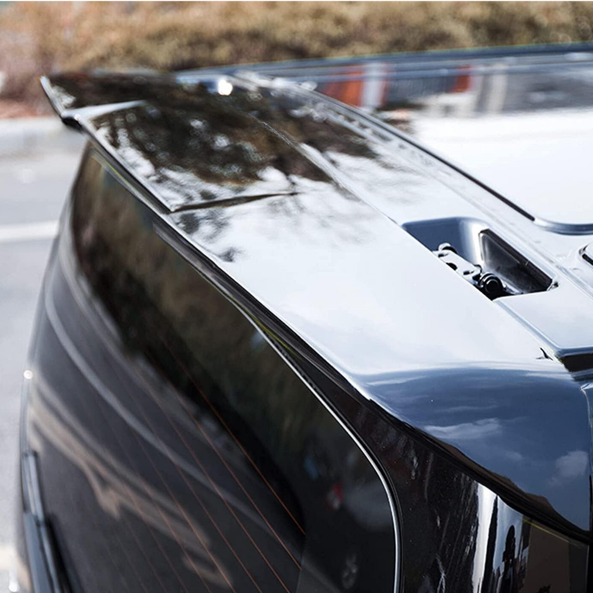 A sleek, glossy car spoiler is installed on the rear of a black vehicle, reflecting trees and sky, situated in an outdoor parking area.