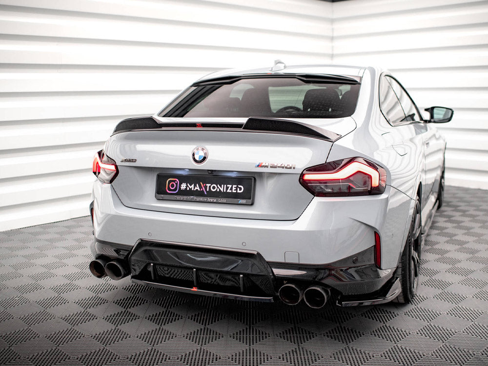 A silver BMW M240i is parked in a white, geometric-patterned room, featuring quad exhausts and elegant taillights. The license plate reads "#MAXTONIZED" with additional text: "Official Premium Partner MAZ 2021."