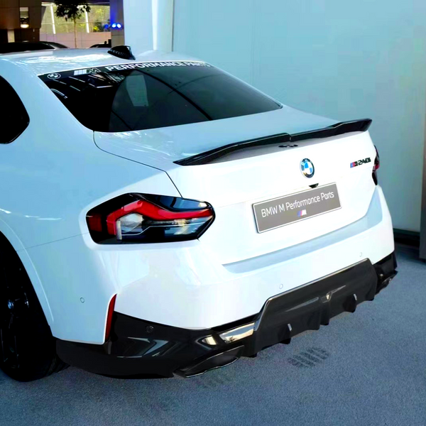 A white BMW car, rear view, features a spoiler and darkened taillights. "M Performance Parts" and "M240i" text is visible. It's displayed indoors on a gray carpeted area.