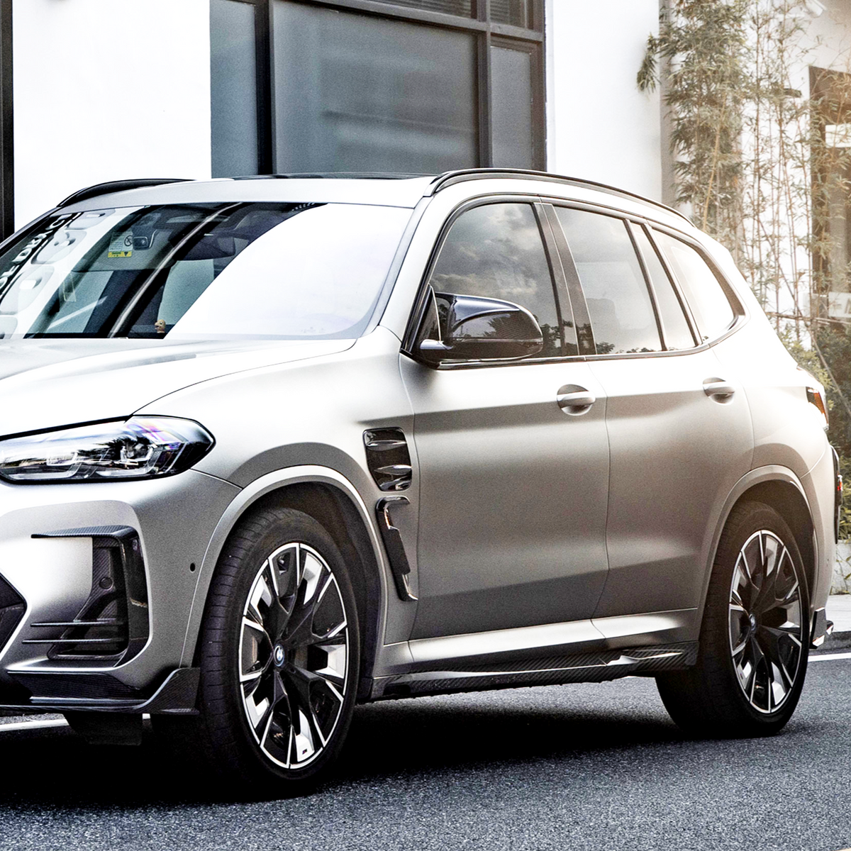 A sleek, silver SUV is parked on a street, featuring stylish alloy wheels and tinted windows, with a white building and small plants visible in the background.