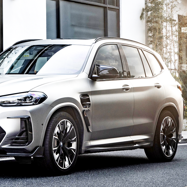 A sleek, silver SUV is parked on a street, featuring stylish alloy wheels and tinted windows, with a white building and small plants visible in the background.
