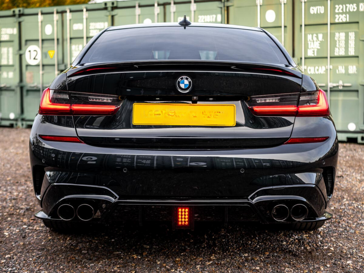 A black BMW with visible exhaust pipes, parked on gravel, is positioned in front of green storage containers. The taillights are illuminated, and the license plate is intentionally blurred out.