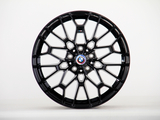 A black alloy car wheel features intricate spoke design, resting stationary, with a BMW logo in the center, set against a plain white background.