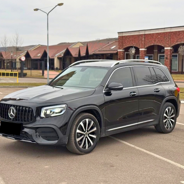 A black SUV is parked in an empty lot, surrounded by brick buildings and leafless trees under a cloudy sky. The vehicle features a prominent front grille and sleek design.