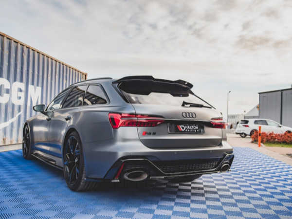 A sleek, silver Audi station wagon parked on a blue checkered surface beside shipping containers and other vehicles in a lot. The license plate reads "D-HAKTON DESIGN."