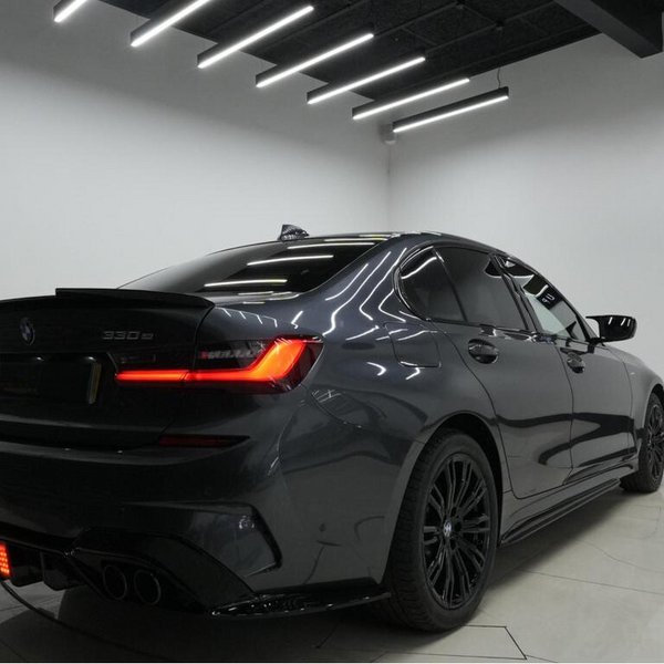 A sleek, dark-colored BMW 330e parked indoors with bright, linear ceiling lights reflecting on its polished surface. The rear of the car features illuminated taillights and visible exhaust pipes.