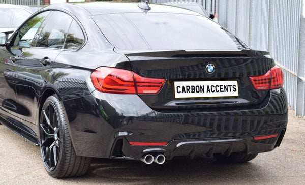 A black BMW car is parked on a street beside a metal fence. The license plate reads "CARBON ACCENTS," and it features dual exhaust pipes and red tail lights.