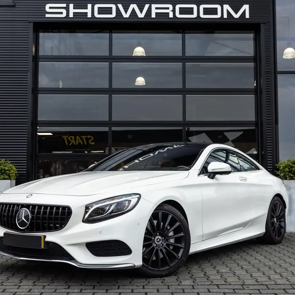 A white Mercedes-Benz car is parked outside a building with "SHOWROOM" written above a large glass door. Two potted plants flank the car on a paved surface.