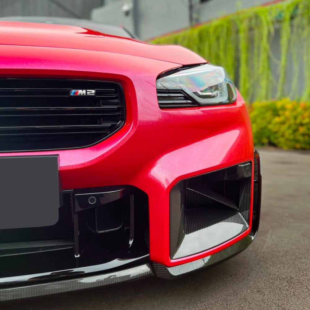 A red car front with a glossy finish displays the "M2" badge on its grille, parked on a paved surface with a wall of greenery in the background.