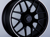 A black alloy wheel with a Mercedes-Benz logo and AMG branding is displayed upright on a dark pedestal against a plain gray background.