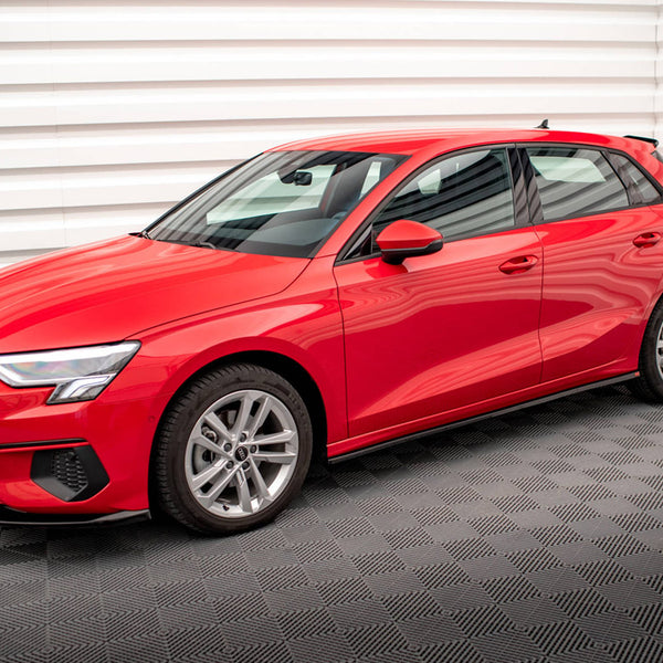 A red hatchback car is parked on a checkered-patterned floor, in a room with white, horizontally ribbed walls. The car features sleek lines and modern design elements.