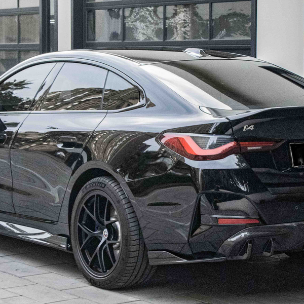 A black BMW i4 parked on a cobblestone driveway outside a modern building with large windows. The car's design features sleek lines and tinted windows, with badges displaying "BMW," "i4," and "M50".