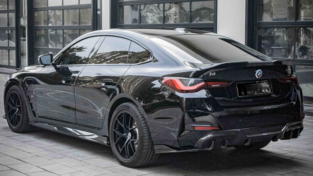 A black BMW i4 parked on a cobblestone driveway outside a modern building with large windows. The car's design features sleek lines and tinted windows, with badges displaying "BMW," "i4," and "M50".