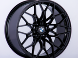 A black, intricately designed BMW alloy wheel is propped against a plain white background, featuring the BMW logo prominently at the center.