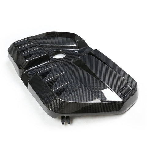 A carbon fiber engine cover with a sleek, angular design lying on a plain white surface. The cover has a central circular opening and raised geometric patterns for added texture.