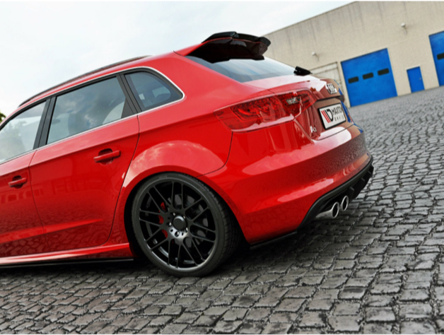 A red hatchback car is parked on a cobblestone surface near a beige building with blue doors. The car's rear features black rims and dual exhaust pipes.