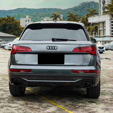 A silver Audi SUV is parked on a concrete surface. In the background, there are trees, hills, and buildings, creating an urban setting with natural surroundings.