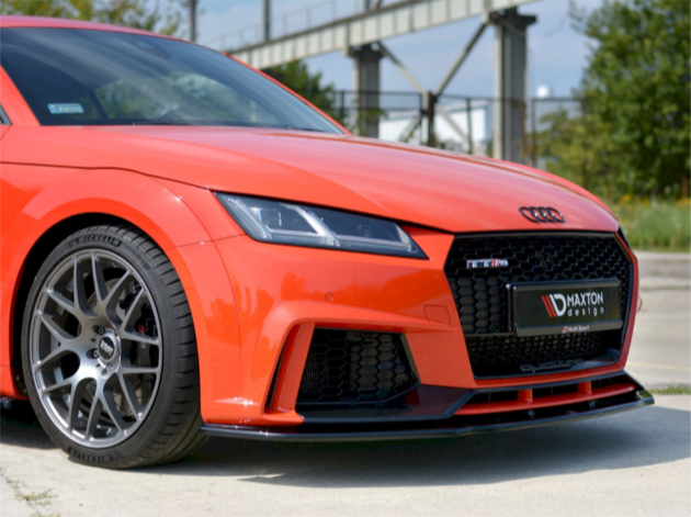 A red sports car with silver alloy wheels is parked on a concrete surface. The front grille displays "Audi" and "Maxton Design" logos. Industrial structures and greenery are in the background.