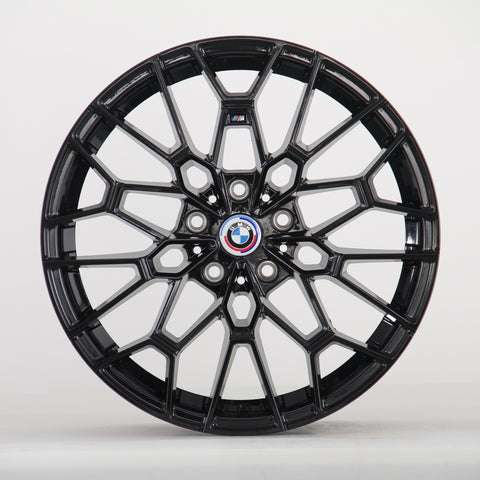 The image shows a black alloy wheel with a complex geometric design, featuring a blue, white, and red BMW logo at the center. The wheel is set against a plain white background.