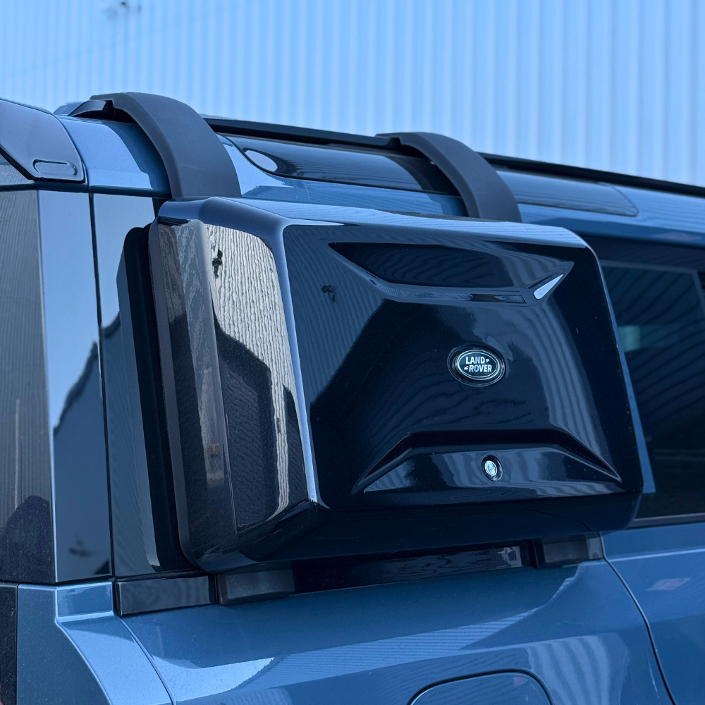 A black, angular box with the "Land Rover" logo is mounted on the rear of a blue vehicle, surrounded by a metallic rooftop in an outdoor setting.