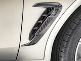 A carbon fiber air vent is attached to the side of a light-colored vehicle, located near the edge of a black tire with visible tread patterns.