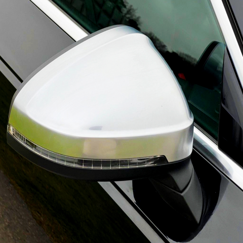 A silver car side mirror is affixed to a black vehicle, reflecting surrounding greenery. The mirror features an integrated turn signal beneath its polished surface.