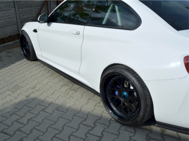 A white car, parked on a gray brick driveway, features black wheels with blue accents and tinted windows, next to a wooden fence.