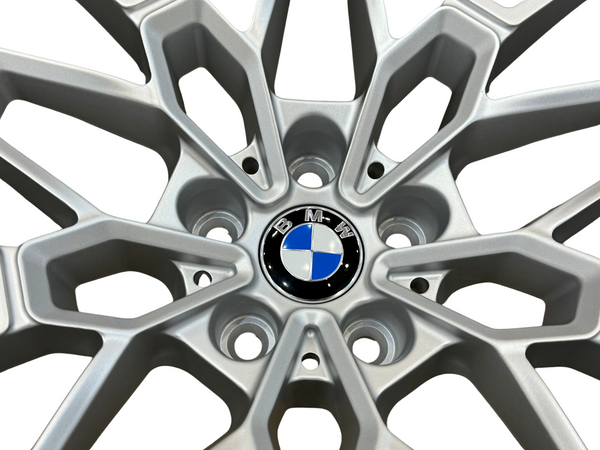 A silver alloy wheel with a geometric design features a central BMW logo, consisting of blue and white quadrants, surrounded by five lug nut holes on a white background.