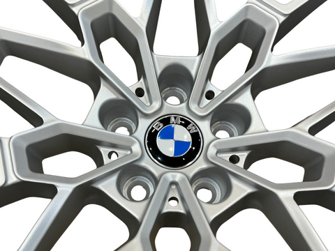 A silver alloy wheel with a geometric design features a central BMW logo, consisting of blue and white quadrants, surrounded by five lug nut holes on a white background.