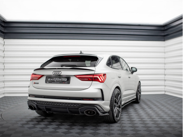 A white Audi car is parked in a modern showroom with geometric black and white walls. The car has sporty black wheels and visible dual exhausts. The license plate reads "#MAXTONIZED."