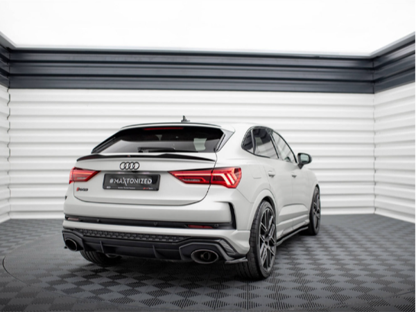 A white Audi car is parked in a modern showroom with geometric black and white walls. The car has sporty black wheels and visible dual exhausts. The license plate reads "#MAXTONIZED."