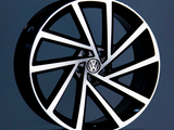 A stylish black and silver alloy wheel rests on a gray platform, featuring the Volkswagen logo at the center, set against a smooth gray background.