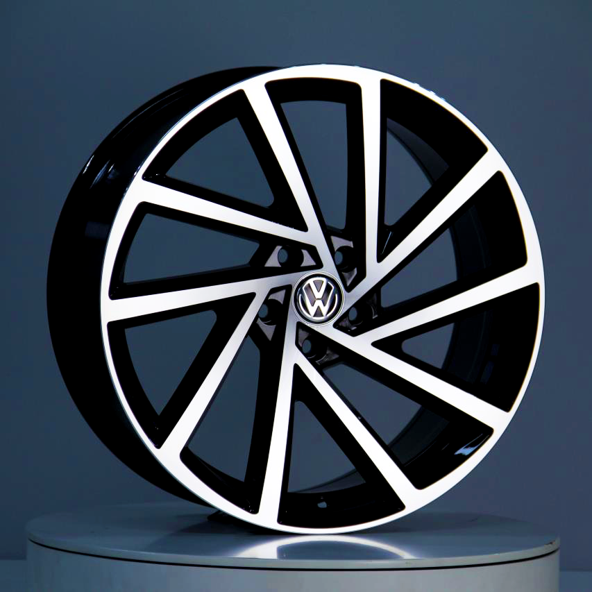 A stylish black and silver alloy wheel rests on a gray platform, featuring the Volkswagen logo at the center, set against a smooth gray background.