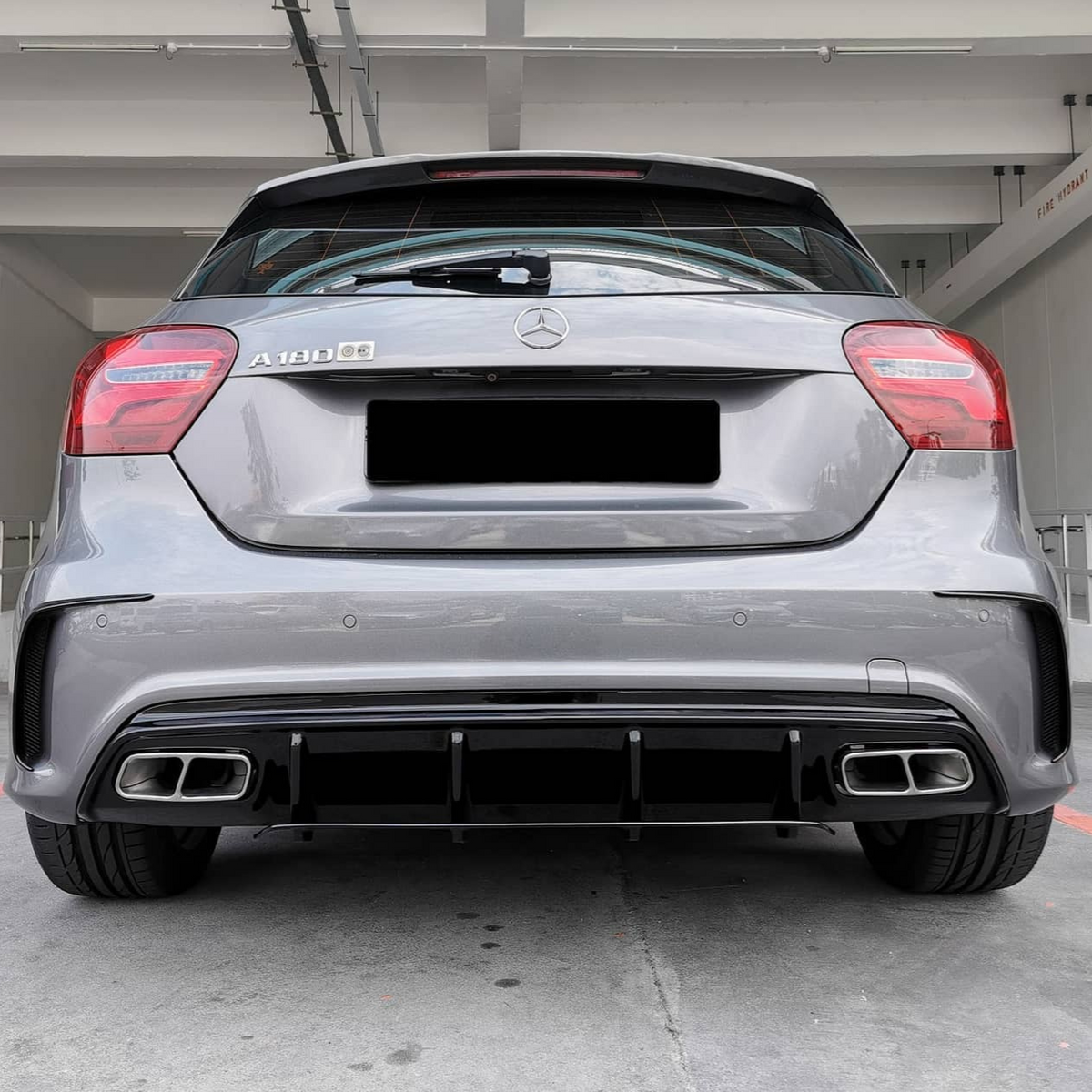 A silver car is parked in a garage, viewed from the rear. It has a visible emblem and dual exhausts. The text reads “A 180” and “FIRE HYDRANT” on a sign.