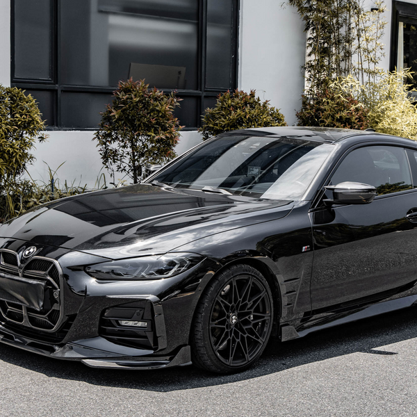 A sleek black sports car is parked on a street, illuminated by sunlight. It's positioned in front of a modern building with large windows, surrounded by decorative shrubs.