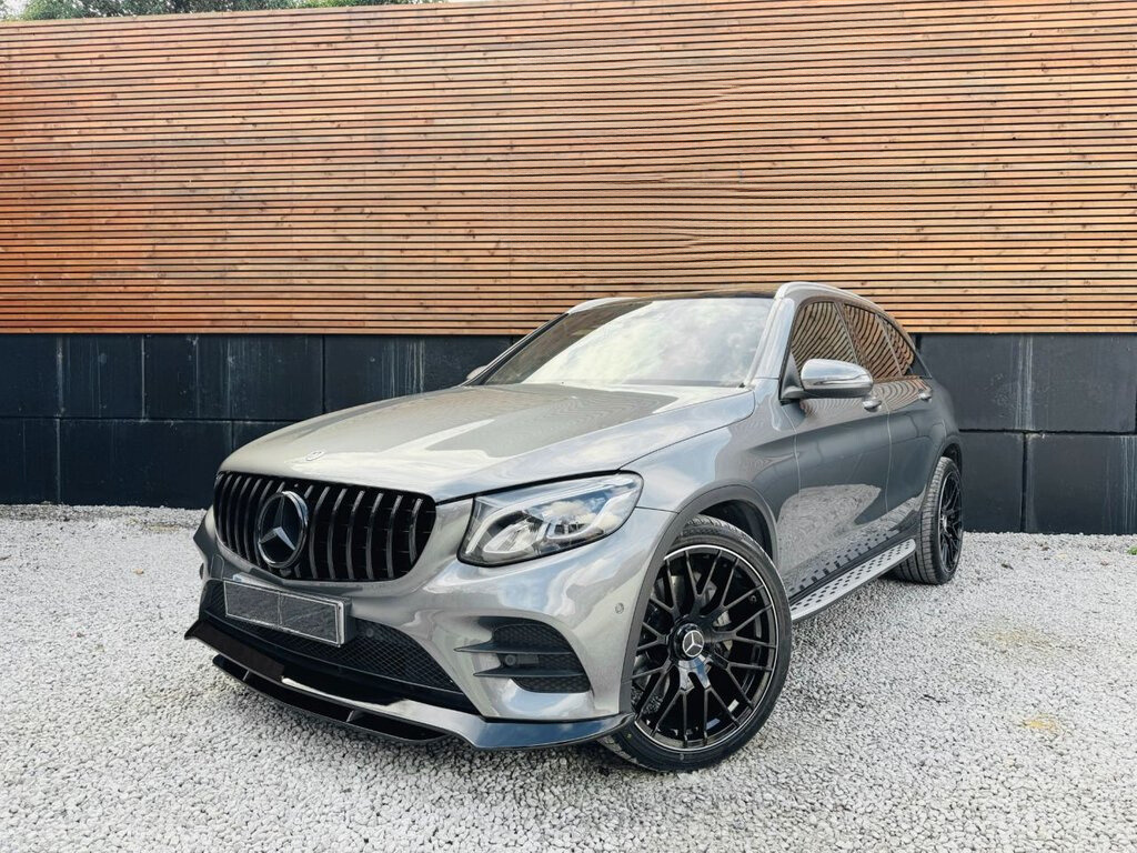 A sleek gray SUV, with large black rims, is parked on gravel. It's positioned in front of a wall with horizontal wooden slats and a black lower section.