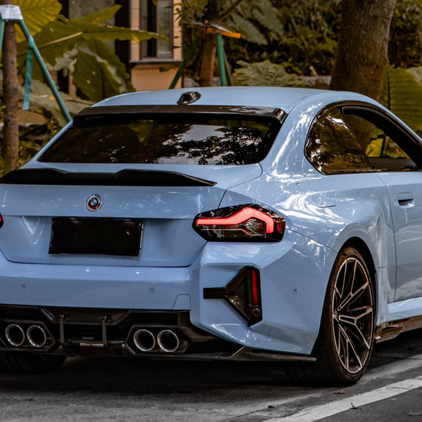 A blue sports car with distinctive tail lights and dual exhausts is parked on a road, surrounded by lush green vegetation and a staircase in the background.