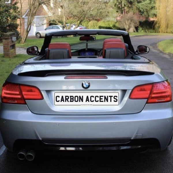 Convertible silver car with red interior parked on a residential street. License plate reads "CARBON ACCENTS." Brick mailbox and house in the background surrounded by trees and grass.