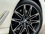 A car wheel with a black and silver alloy rim, featuring the BMW logo, is stationary on a road. The vehicle’s white body and a nearby leaf are visible.