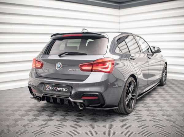 A grey BMW car is parked in a showroom with patterned flooring and white paneled walls. The car has "F21 M" and "maxtonized" written on the back.