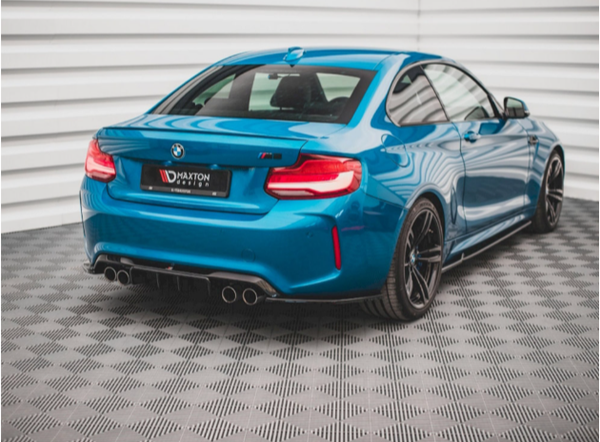 A blue BMW sports car is parked in a showroom with a textured gray floor and white walls. The vehicle features quad exhaust pipes and the text "BMW," "M2," and "MAXTON Design."