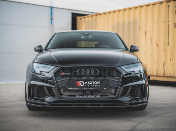 A black Audi car is parked on concrete flooring, facing forward. The license plate reads "MAXTON DESIGN." A large shipping container and a gray building are in the background.