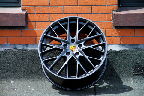 A black alloy wheel with a yellow Porsche emblem, leaning against an orange brick wall, casting a shadow on a textured ground surface.