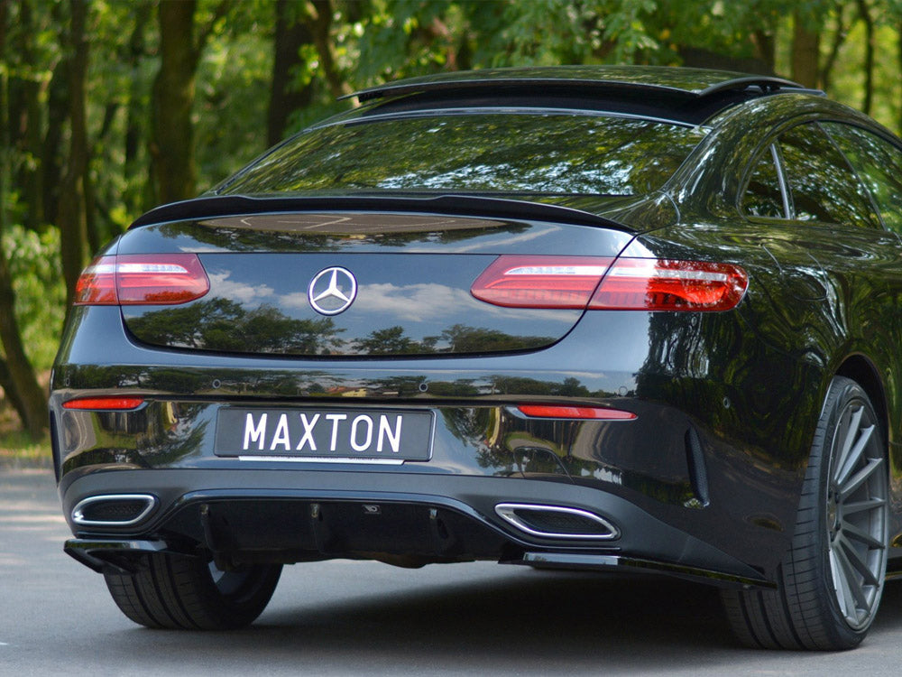 A black car with a logo on the rear and exhaust openings, parked on a road. The license plate reads "MAXTON." Trees are visible in the background.