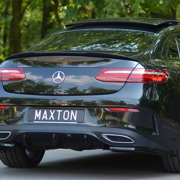 A black car with a logo on the rear and exhaust openings, parked on a road. The license plate reads "MAXTON." Trees are visible in the background.