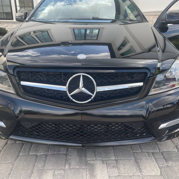 A black Mercedes-Benz car is parked on a stone driveway outside a building. The car's front grill and emblem are prominently visible, with the driver's door slightly open.