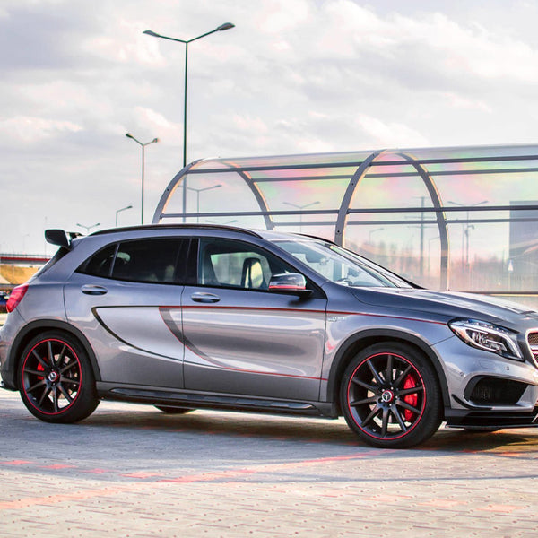 A silver hatchback car with red-accented black wheels is parked on a paved lot. The vehicle features a sleek design. The background has a glass-covered walkway and lamp posts.